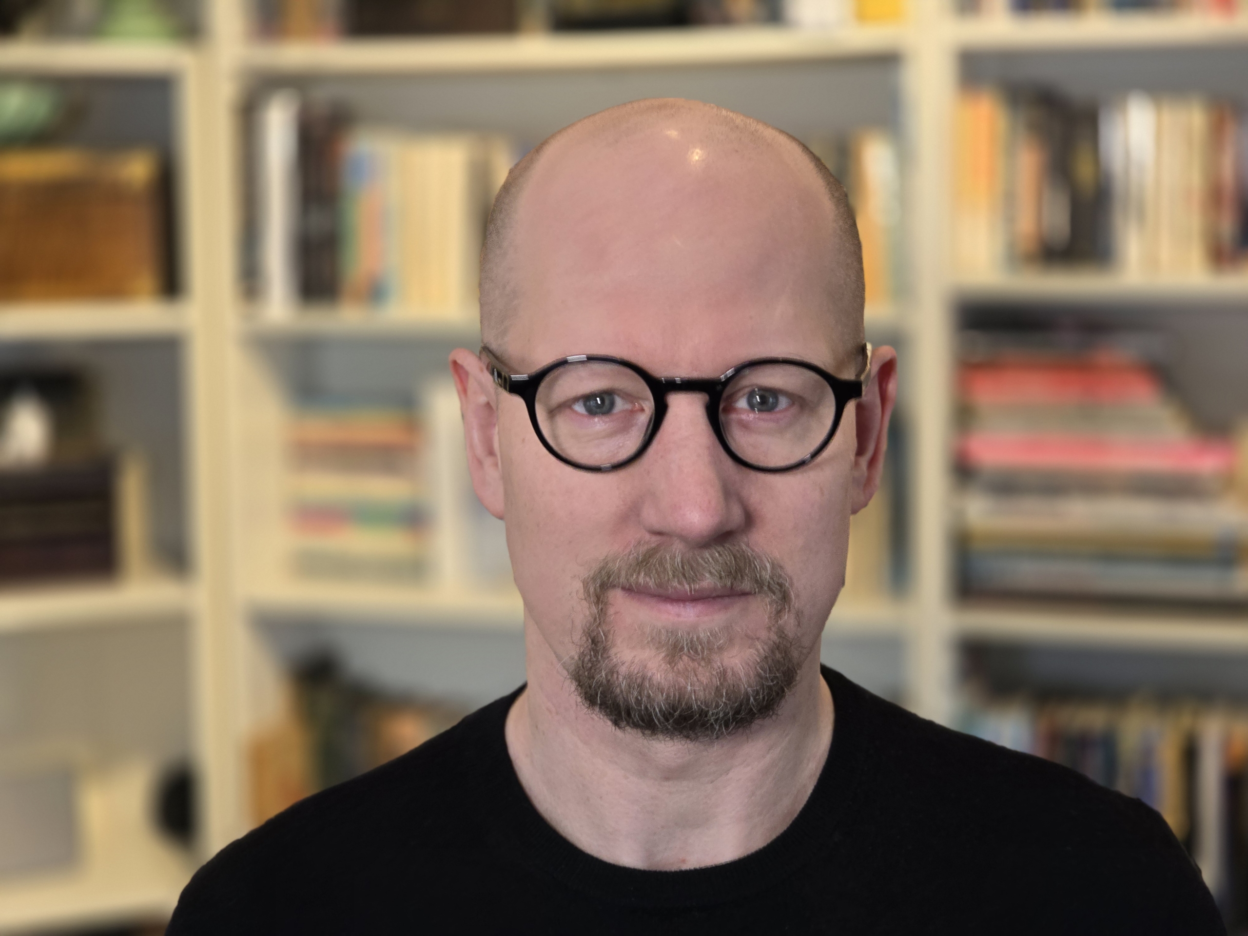 En person med glasögon och svart tröja står framför en bokhylla