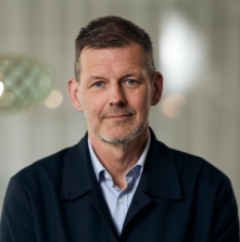 En person med kort hår och skäggstubb, klädd i en mörk jacka och ljusblå skjorta