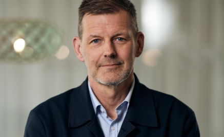 En person med kort hår och skäggstubb, klädd i en mörk jacka och ljusblå skjorta