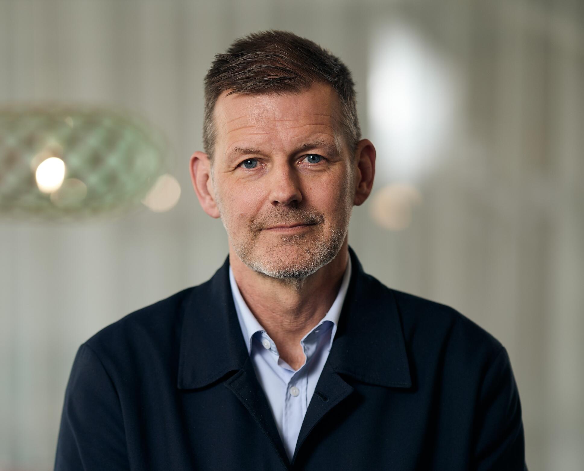 En person med kort hår och skäggstubb, klädd i en mörk jacka och ljusblå skjorta